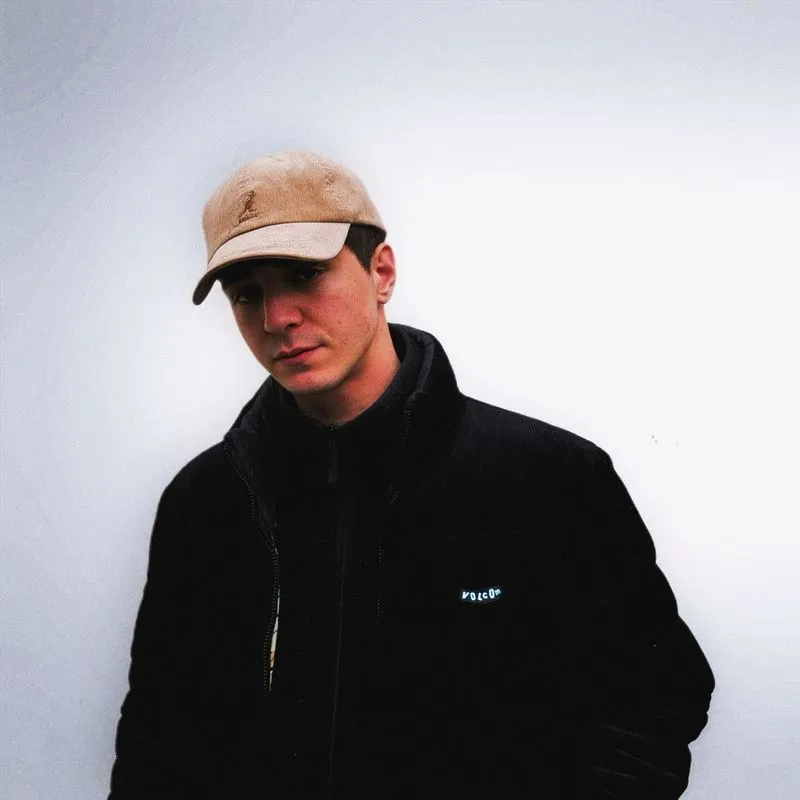 Automhate wearing a beige cap and dark jacket against a pale studio background.