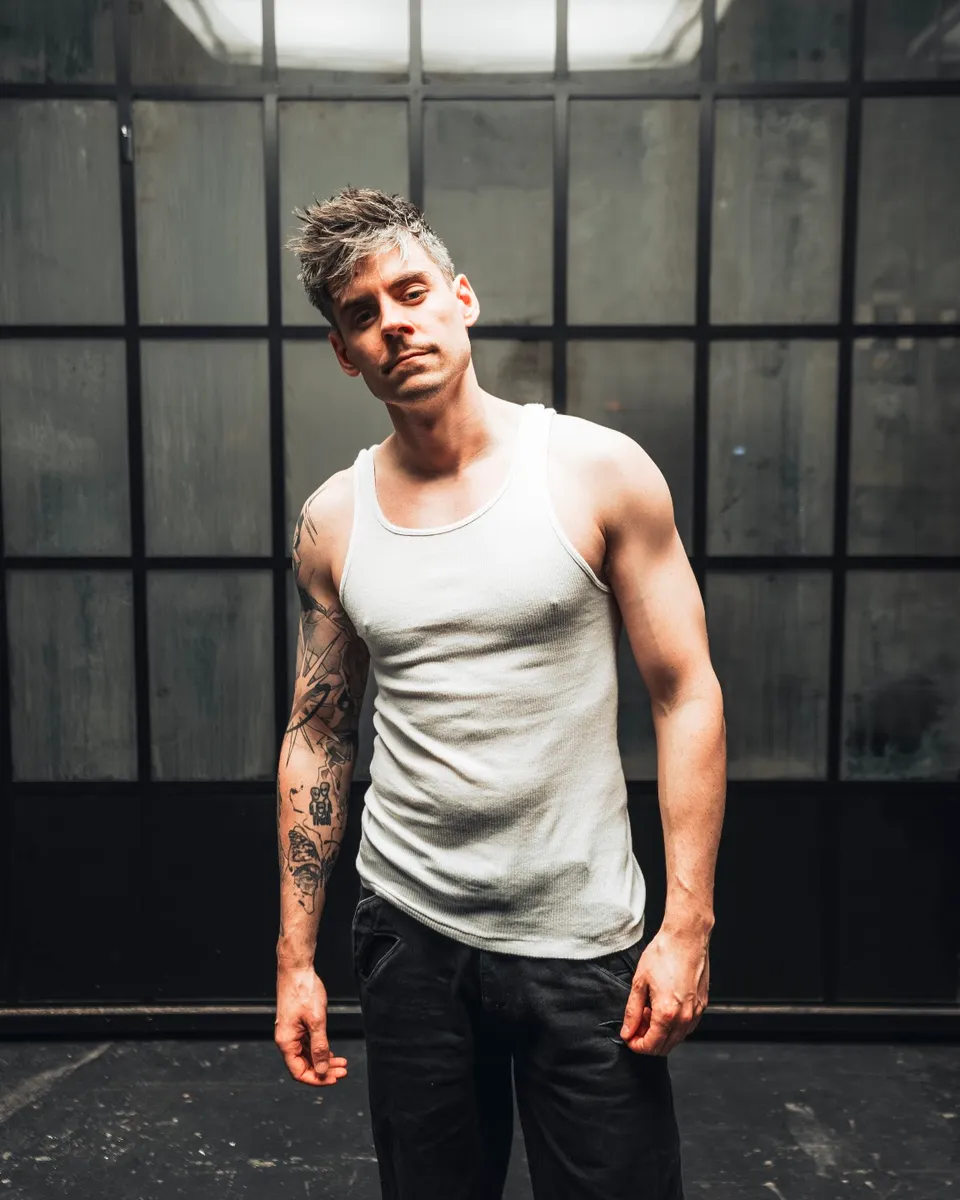 Fairlane in a white vest standing in front of tall studio windows.