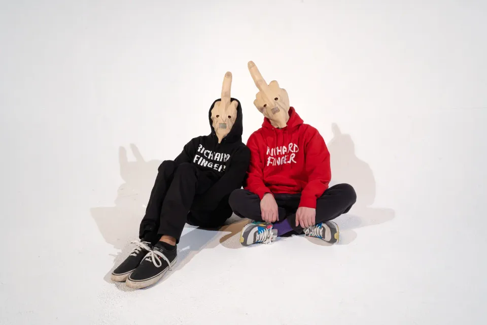 The Richard Finger duo seated together wearing finger-shaped masks and black and red branded hoodies.