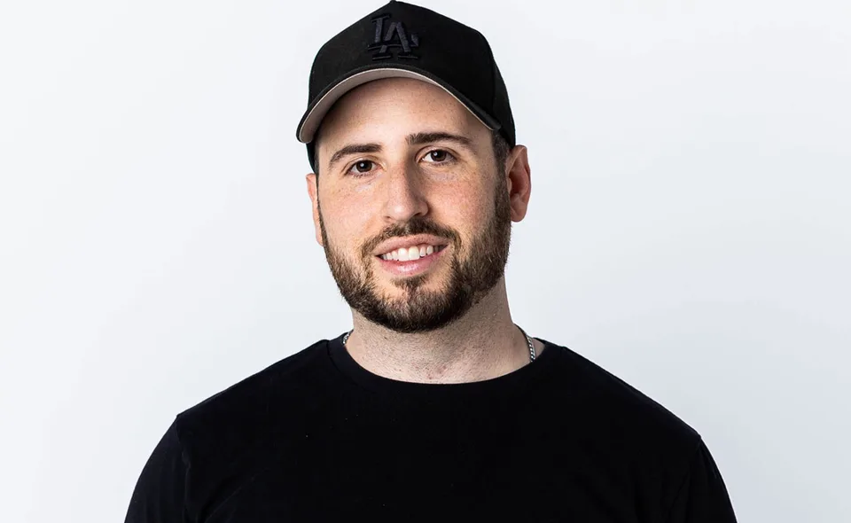 Wooli smiling in a black cap and black shirt against a pale background.